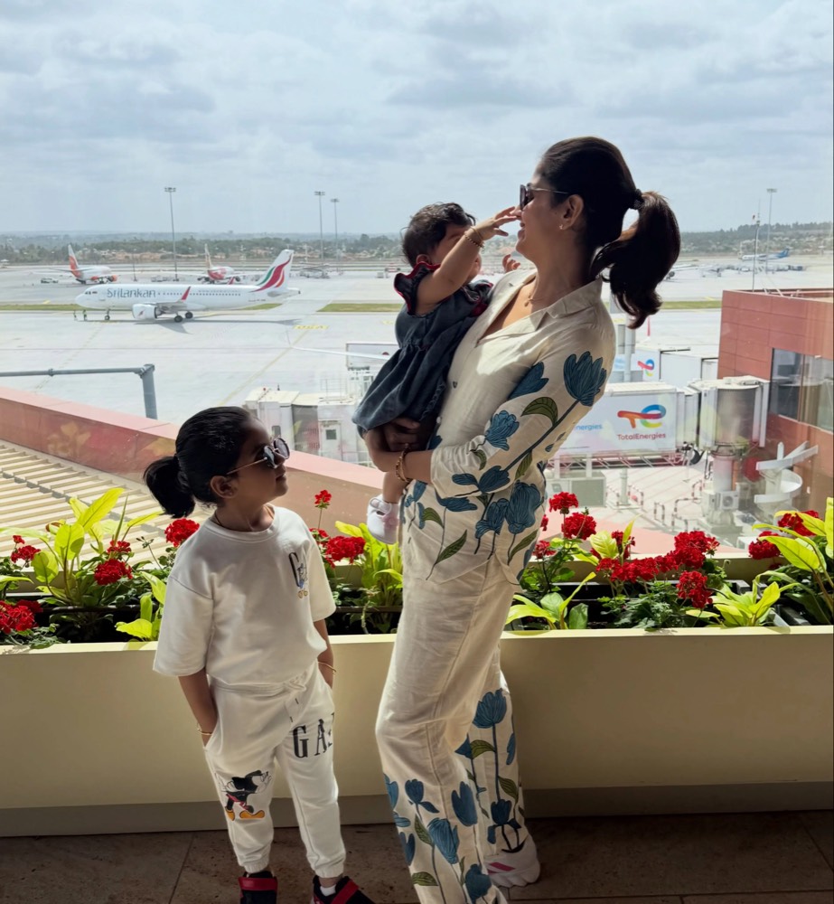 Chaithra Rai lifting her baby daughter at an airport window while her elder daughter watches, aircraft on the apron behind