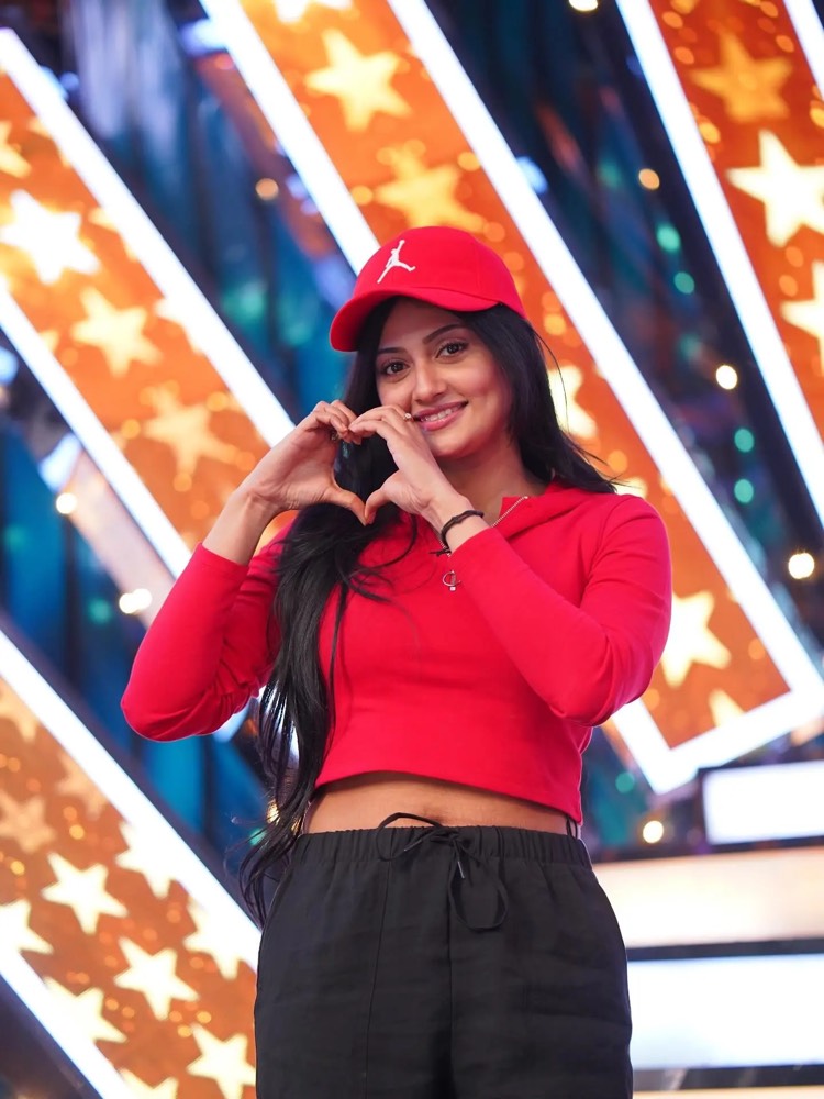 Debjani Modak in a red cap and red top making a heart with her hands on a lit stage set