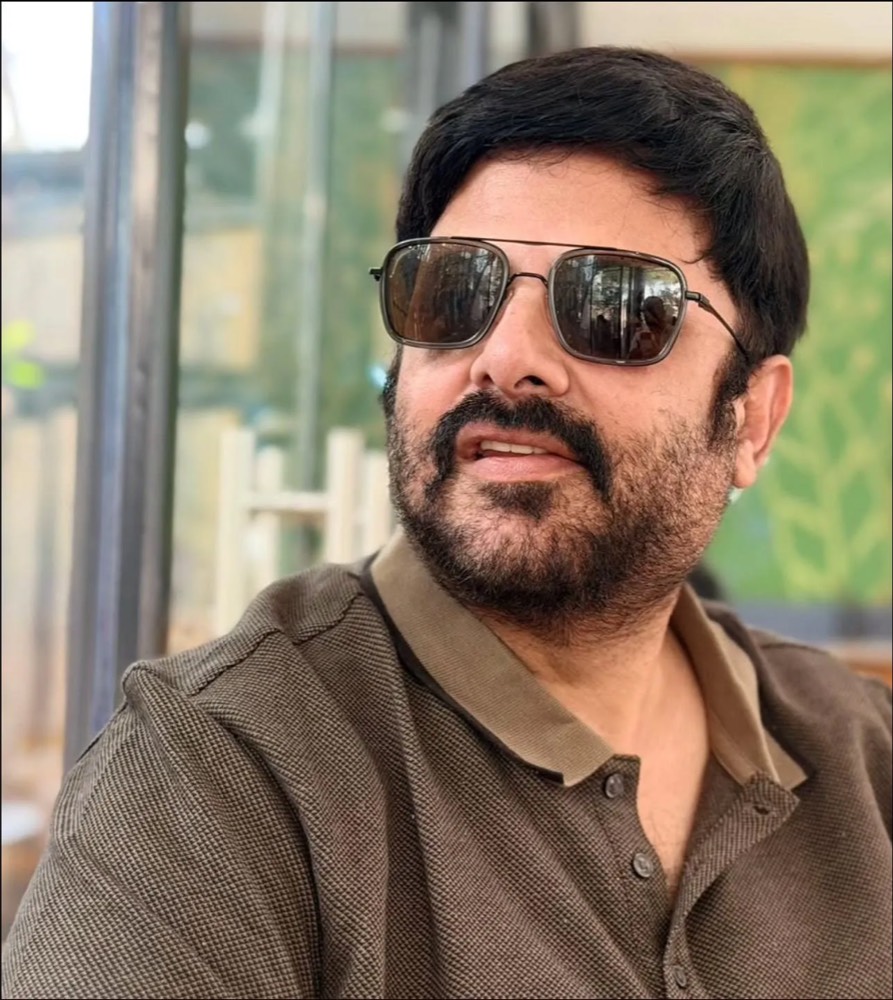 Krishnudu in sunglasses and an olive polo shirt, mid-sentence, sitting by a window with a green wall behind him