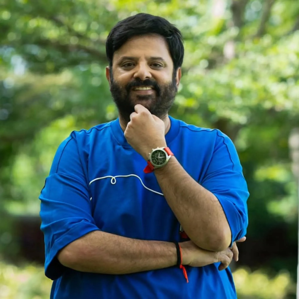 Krishnudu standing outdoors in a blue T-shirt with his arms folded and a hand at his chin, green foliage blurred behind him
