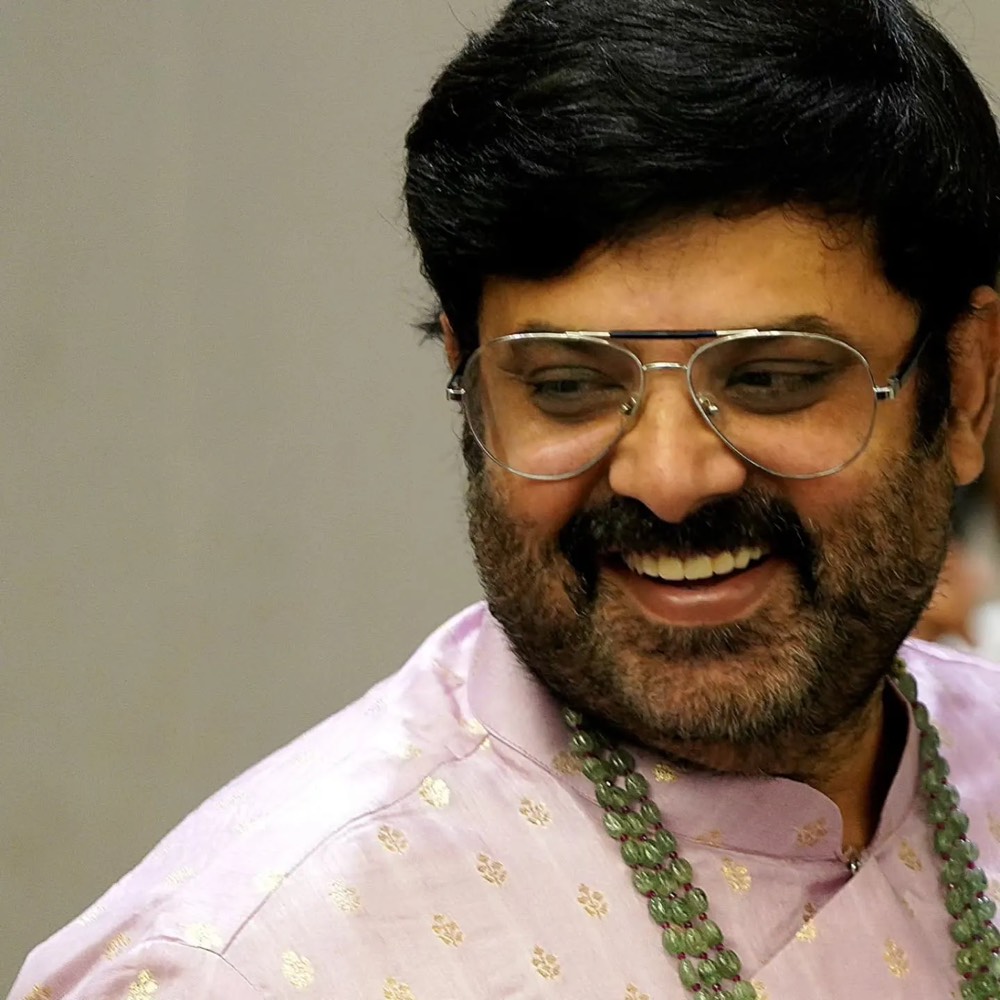 Krishnudu laughing, in a pale pink kurta with gold motifs and a string of green beads, wearing aviator glasses