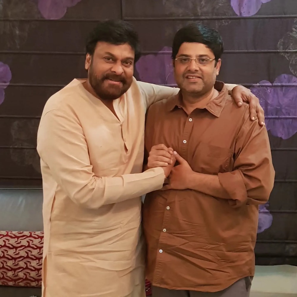 Krishnudu in a brown shirt standing arm in arm with the actor Chiranjeevi, who is in a cream kurta, in front of a dark panelled wall
