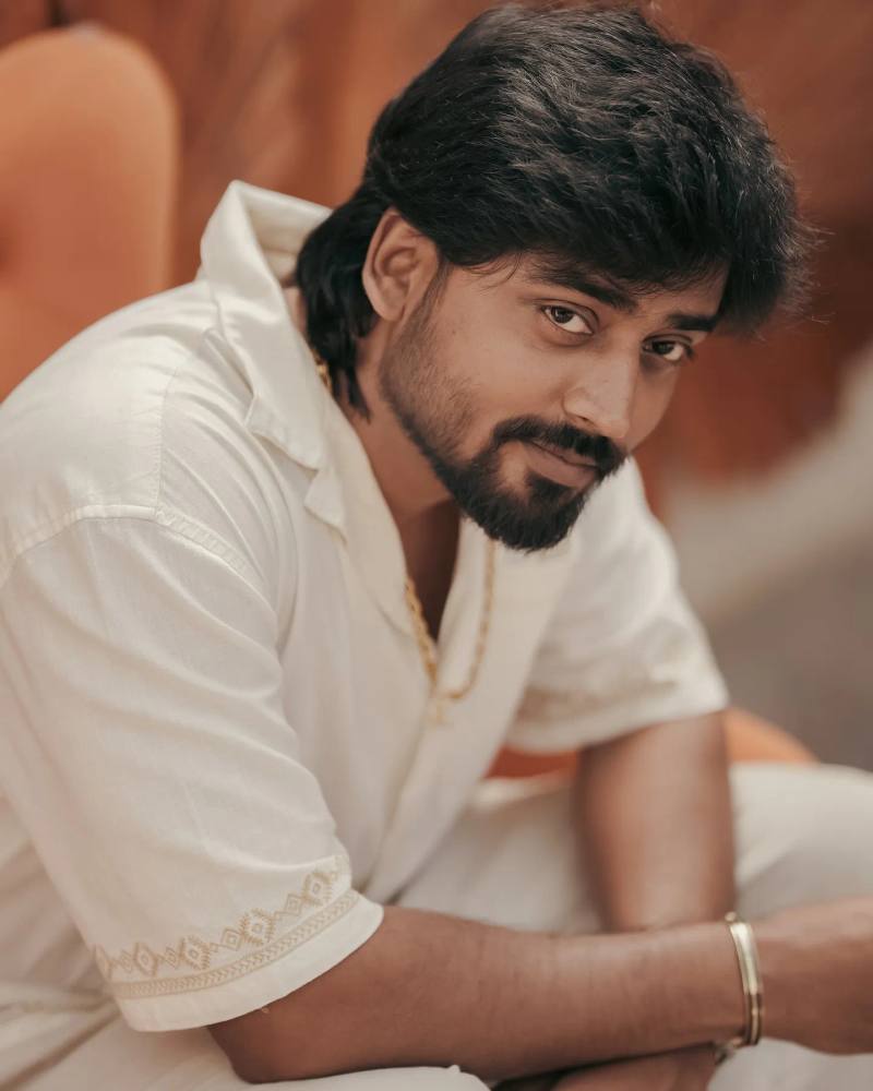 Mukesh Gowda in a white shirt, seated and leaning forward, in warm light