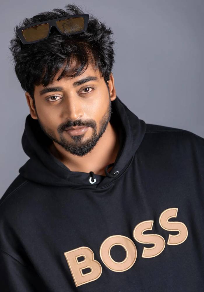 Mukesh Gowda in a black hoodie with sunglasses pushed up into his hair, against a grey backdrop