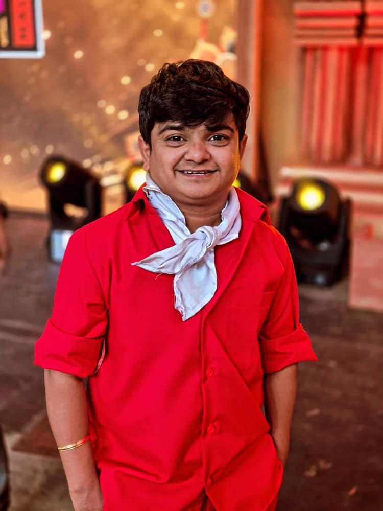 Naresh Jabardasth in a red shirt with a white scarf knotted at his neck, stage lights behind him