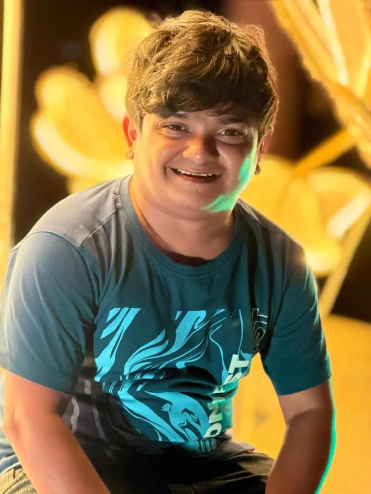 Naresh Jabardasth sitting and laughing in a teal printed T-shirt, warm out-of-focus lights behind him