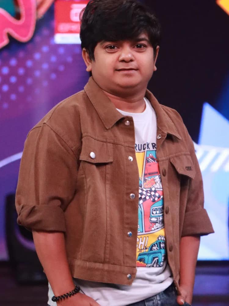 Naresh Jabardasth in a brown denim jacket over a printed T-shirt, standing on a studio floor
