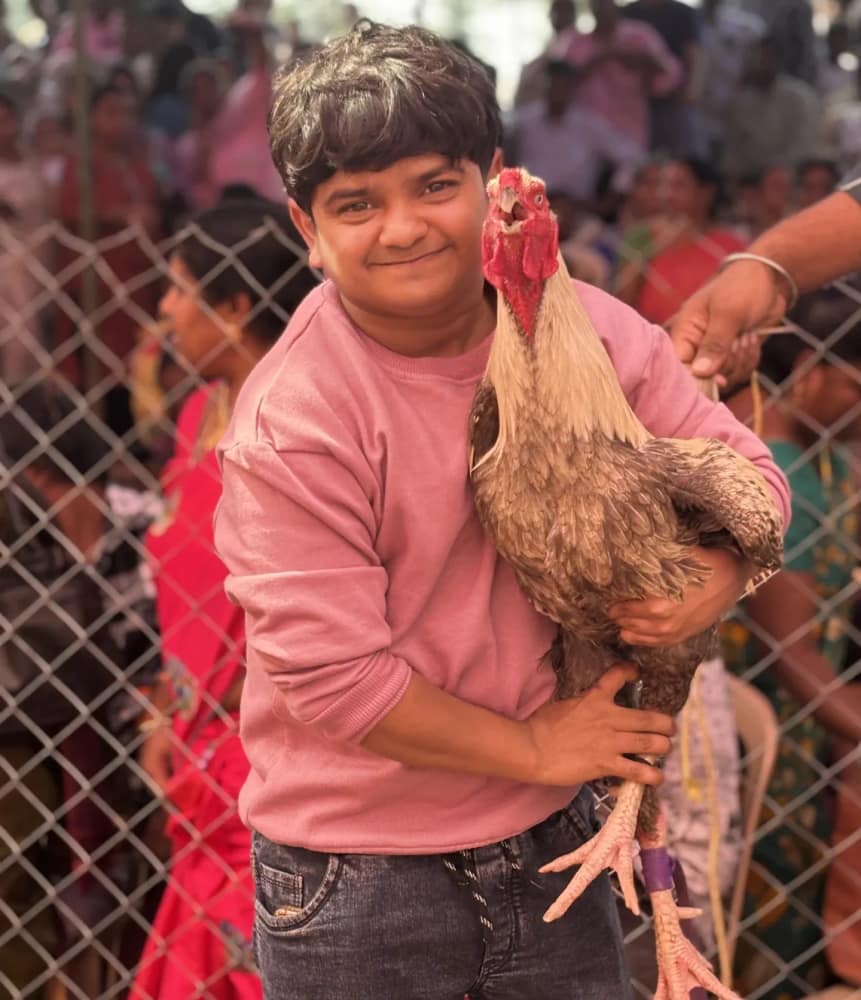 Naresh Jabardasth in a pink sweatshirt holding a rooster, with a crowd behind a wire fence