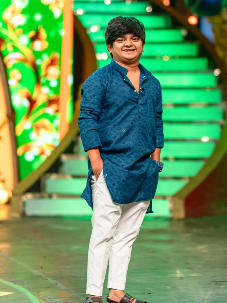 Naresh Jabardasth standing on a television set in a blue embroidered kurta and white trousers, hands in his pockets