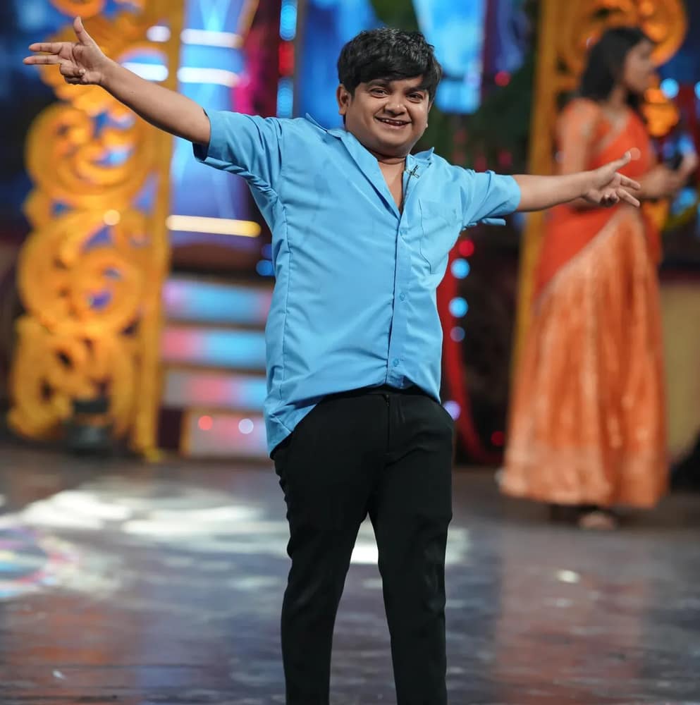 Naresh Jabardasth mid-move on a stage in a light blue shirt and black trousers, both arms flung out