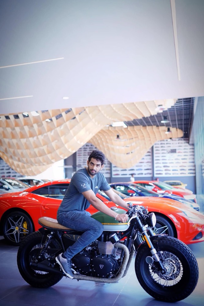 Sudheer Reddy sitting on a Triumph motorcycle in a showroom with sports cars parked behind him