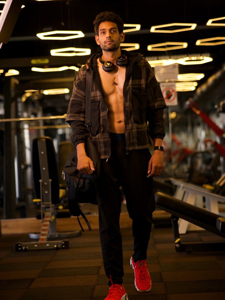 Sudheer Reddy walking across a gym floor in an open checked shirt with headphones round his neck and a kit bag on his shoulder