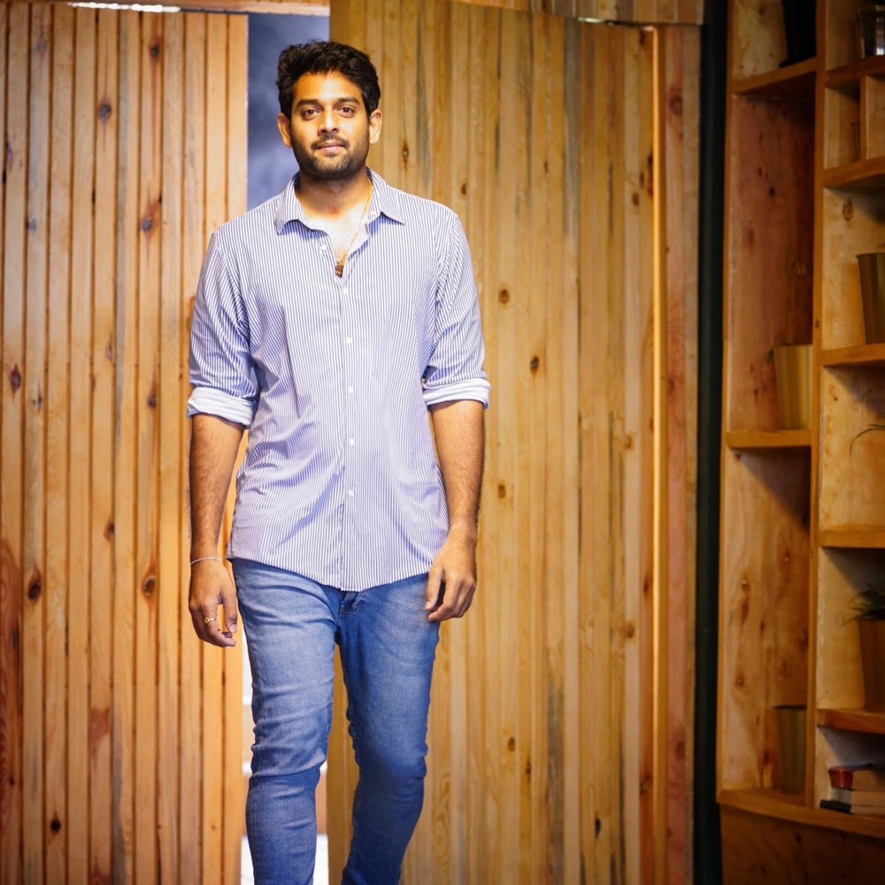 Sudheer Reddy walking through a wood-panelled room in a blue striped shirt and jeans