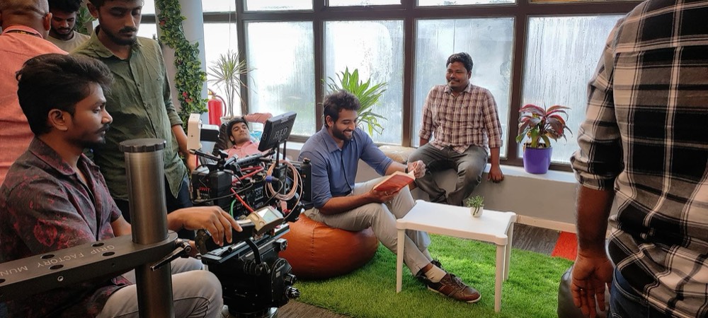 Sudheer Reddy sitting on an orange bean bag reading from a notebook while a camera crew films him and a guest sits opposite