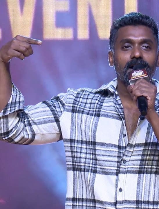Temper Vamsi on stage in a black and white checked shirt, pointing at himself while speaking into a microphone