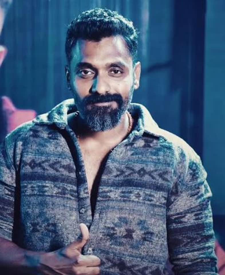Temper Vamsi in a patterned grey overshirt, half smiling, in a scene lit blue