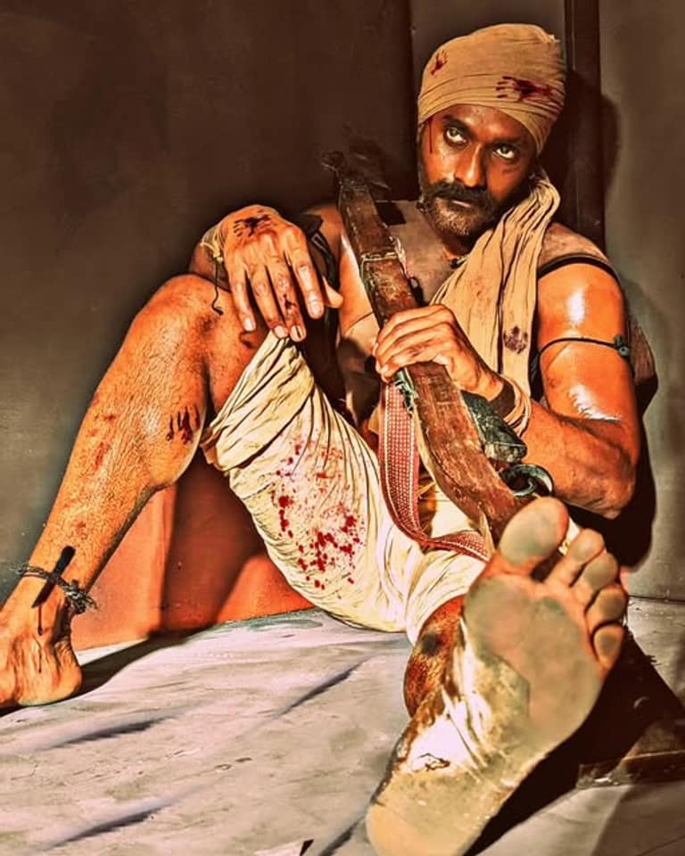 Temper Vamsi in character, bloodied, wearing a headcloth and holding an axe against a wall