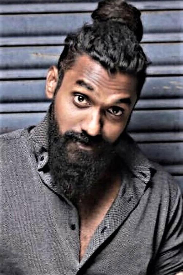 Temper Vamsi with a full beard and his hair in a top knot, leaning against a metal shutter